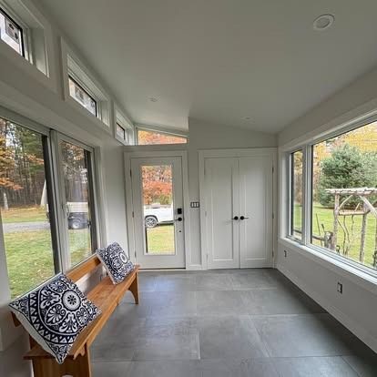 A sunroom with gray tile flooring, a wooden bench with patterned pillows, white double doors, and windows overlooking woods.