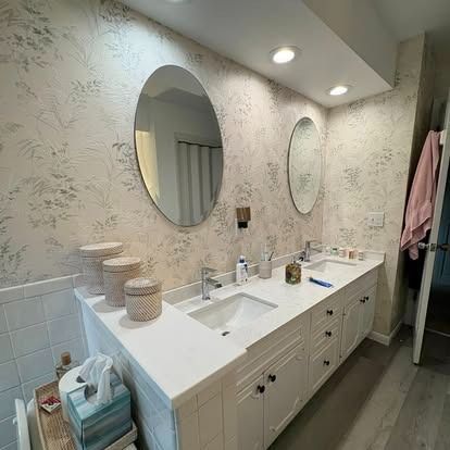 A double-sink bathroom vanity with white cabinets, light countertops, two oval mirrors, and light botanical wallpaper.