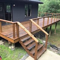 A wooden deck with cable railings and stairs leading to a dark gray house in a wooded area.