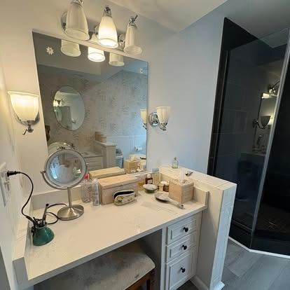 A white bathroom vanity with a mirror, vanity light, makeup mirror, and drawers, next to a glass-enclosed shower.