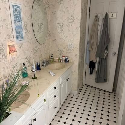 A bathroom vanity with floral wallpaper, a round mirror, white cabinets, and robes hanging on the door.