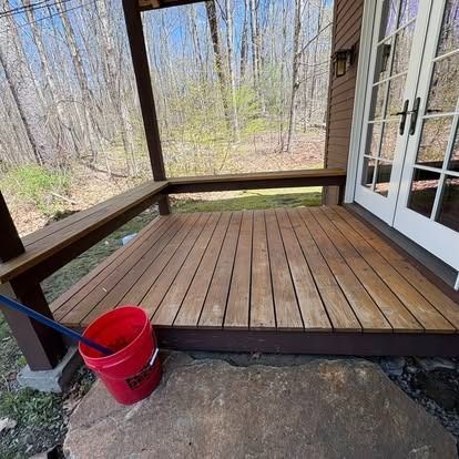 A wooden deck with a built-in bench under a covered porch, set against a backdrop of trees, with a red bucket in front.