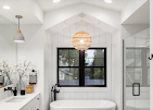 A modern bathroom with a freestanding tub, dark-framed window, woven pendant light, and white cabinetry.