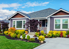 A single-story suburban house with dark gray siding, white trim, and a manicured front garden under a partly cloudy sky.