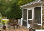 A wooden deck with a bench and a dining set next to a white-framed screened porch attached to a brick house.