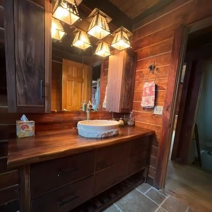 A rustic bathroom vanity with a live-edge wood countertop, stone vessel sink, and warm wooden walls with lantern lights.