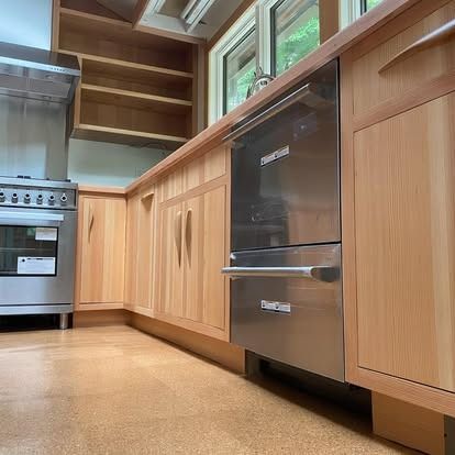 Light wood kitchen cabinets and countertops with a stainless steel dishwasher and oven in a sunlit room with cork floors.
