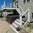 A multi-level composite deck with white railings and stairs leads to a stone patio featuring a hot tub under the deck.
