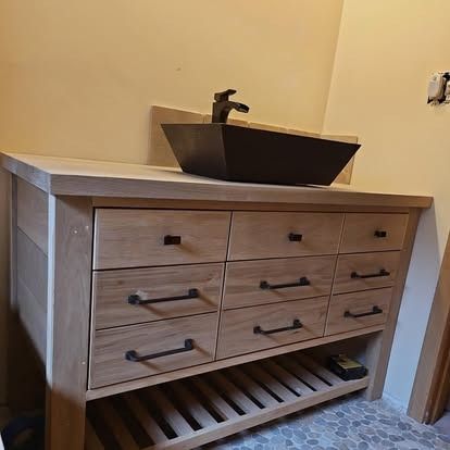 A light-toned wooden vanity with nine drawers and an open slatted shelf, topped with a dark rectangular basin sink.