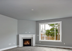 A gray room featuring a white corner fireplace and a large window looking out onto a fenced yard with trees.