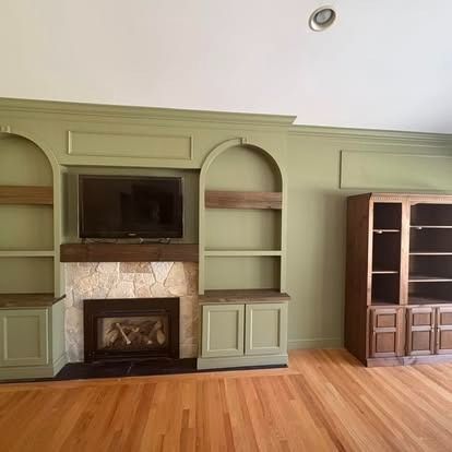A room with sage green built-in shelving, a stone fireplace with a mounted television, and a wooden cabinet on a wood floor.