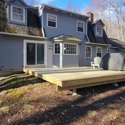 A new light wood deck extends from a blue two-story house with a white chair and covered grill on a wooded lot.