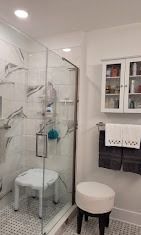 A modern bathroom with a glass-enclosed shower containing a white shower chair, next to a small white stool and wall cabinet.