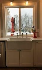 A white farmhouse sink sits under a bright window looking out on a snowy, wooded yard.