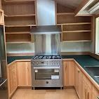 A kitchen with natural wood cabinets, open shelves, green countertops, and a stainless steel stove with a matching hood.