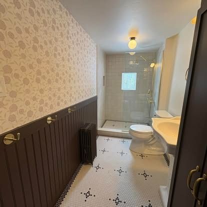 A bathroom with floral wallpaper, dark brown wainscoting, a pedestal sink, and a tiled shower stall with a glass door.