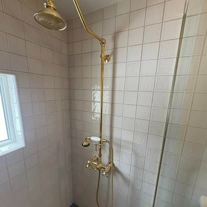 A brass shower head and fixture set mounted on a wall with white square tiles next to a small window.