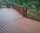 A wide outdoor deck with brown composite planks, matching railings, and a view of green trees in the background.