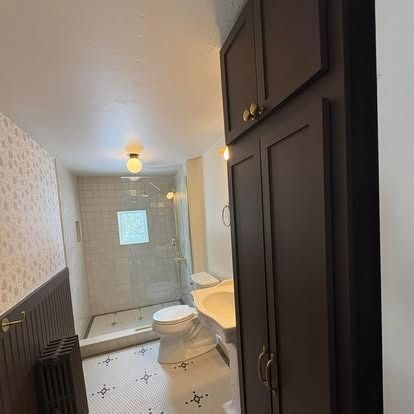 Bathroom with dark brown cabinetry, a pedestal sink, white toilet, and a glass-enclosed shower on tiled floors.