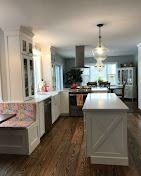 Modern white kitchen with a center island, wooden floors, built-in dining nook, and a prominent glass pendant light.