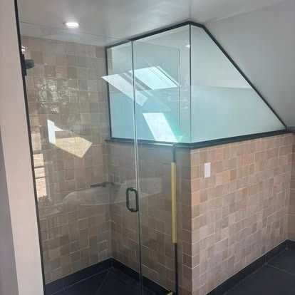 A walk-in shower with beige square wall tiles, a glass door, and an angled glass partition under a sloped ceiling.