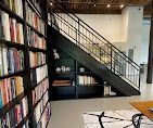 Black bookshelves filled with books line a wall next to a staircase with a black metal railing and storage shelves below.