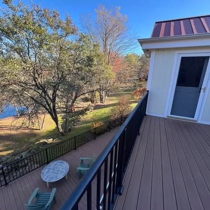 Elevated wooden deck with black railings overlooking a lawn, trees, a round table, and two chairs on a sunny day.