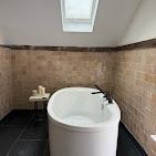 A freestanding oval white bathtub in a bathroom with beige tiled walls, dark stone floors, and a roof skylight.