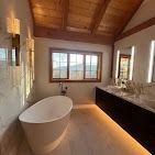 Modern bathroom with a freestanding white tub, dark vanity, illuminated mirrors, and a wood-beamed ceiling.