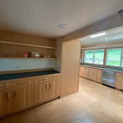 Kitchen with light wood cabinets, black countertops, open shelving, and a large window looking out to a grassy field.