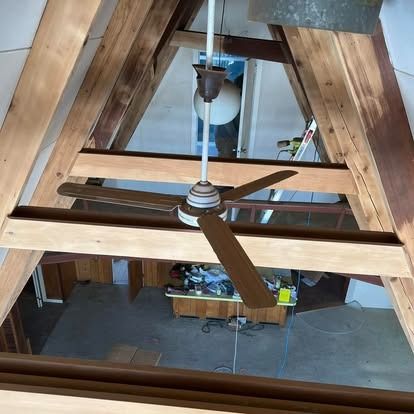 An A-frame room viewed from above, featuring a wooden ceiling structure, a central ceiling fan, and a lower work area.