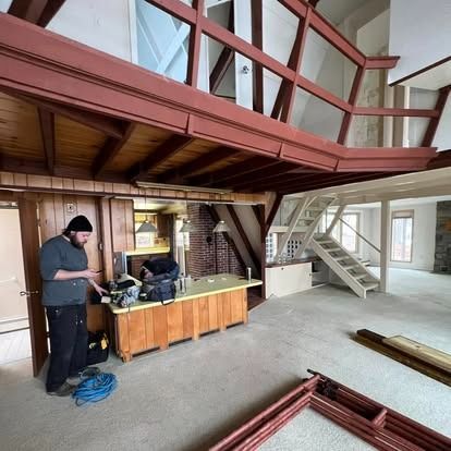 A person in dark clothes stands in a rustic room under renovation with wood beams and a staircase leading to a loft.
