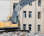 A yellow excavator sits in front of a multi-story building, with a pile of rubble in the foreground.
