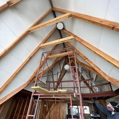 A person in a respirator mask stands on scaffolding below high, vaulted wooden beams and white ceiling panels.