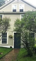 Two-story light-beige house with a dark door, windows, and a brick path leading to the entrance, framed by trees.