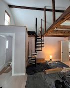 A black metal spiral staircase connects the main floor to a loft area in a room with light grey walls and wooden beams.