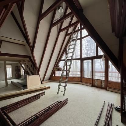 An A-frame house interior under renovation with a tall ladder, white walls, exposed wooden beams, and a large window wall.