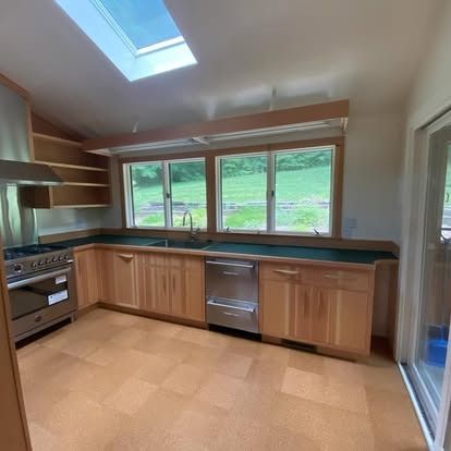 A bright, modern kitchen with light wood cabinets, dark countertops, a stainless steel stove, and a skylight above.