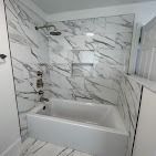 A modern bathroom bathtub and shower with white and grey marble-patterned tile walls and built-in shelving.