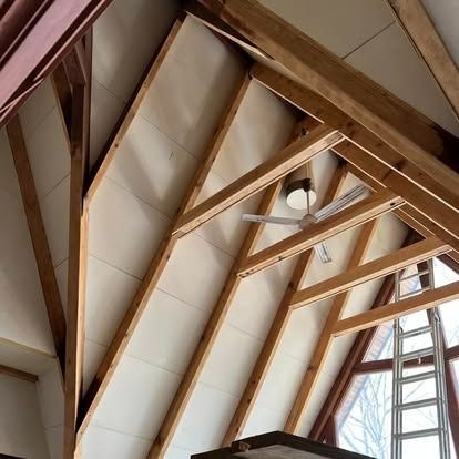 A low-angle view of an A-frame interior featuring wooden beams, white ceiling panels, a ceiling fan, and a metal ladder.