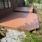 A two-level wooden deck with reddish-brown planks extends from a house to a stone patio.