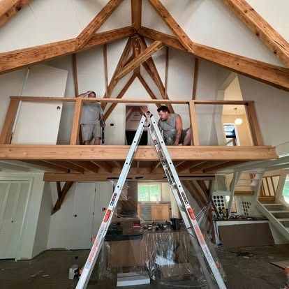 Two workers in a room with a wooden vaulted ceiling build a railing on a loft level, viewed past an open stepladder.