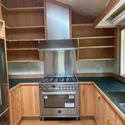 Stainless steel range and vent hood in a kitchen with natural wood cabinets, open shelves, and dark green countertops.