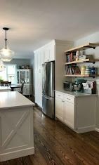 A bright kitchen with white cabinetry, a stainless steel refrigerator, open wood shelving, and dark wood flooring.