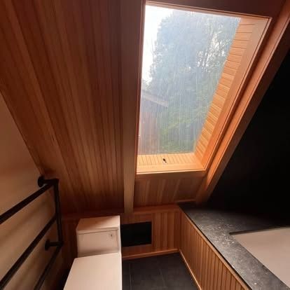 A modern, wood-paneled bathroom featuring a slanted skylight above a bathtub, with a toilet nearby and dark tiled floors.