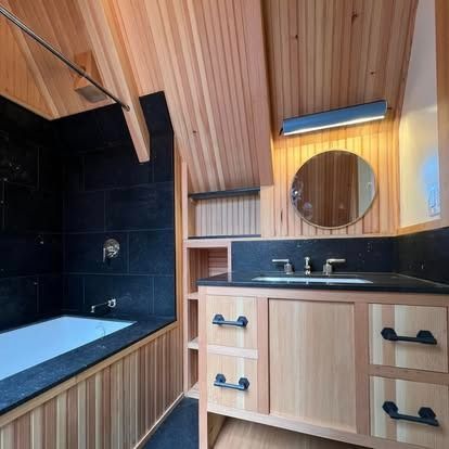 A modern bathroom features light wood paneling, a dark tiled bathtub area, and a vanity with black hardware and a mirror.