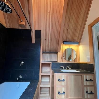 Modern bathroom with a black-tiled shower, white tub, light wood slatted ceiling, shelving, and a wood vanity with a mirror.