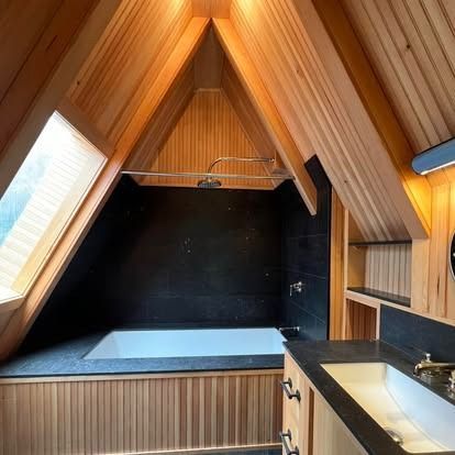 A modern, A-frame bathroom featuring a white soaking tub, black tile walls, and light wood paneled ceilings and cabinets.