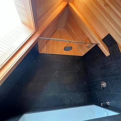 A bathtub set into a wall of dark slate tiles beneath a sloped, natural wood paneled ceiling with a rainfall showerhead.