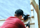 A person wearing a red shirt, black hat, and work gloves uses a tool to install siding on the exterior of a building.